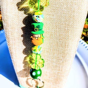 Könnte beinhalten: Ein St. Patrick's Day-Themen-Lanyard mit grünen Perlen, Kleeblatt-Anhängern und einem Kobold-Anhänger. Das Lanyard wird auf einer mit Jute bedeckten Büste präsentiert. Das Lanyard hat eine grüne Kordel und einen Metallverschluss.