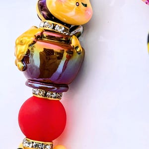 Puede incluir: Un dije caprichoso con una abeja amarilla, un tarro de miel, una esfera roja y un oso sonriente. La abeja y el oso son amarillos, el tarro de miel es iridiscente y la esfera es roja. Todos los elementos están adornados con pedrería.