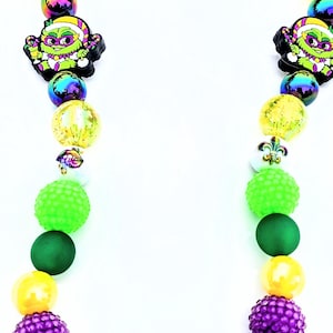 May include: A Mardi Gras necklace featuring a variety of beads in yellow, green, purple, and iridescent colors. The necklace includes decorative mask charms and other festive elements.
