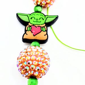 Puede incluir: Un accesorio de cuentas con un dije de Grogu verde sosteniendo un corazón, rodeado de cuentas cubiertas de cristales iridiscentes y cuentas de color verde y melocotón. El accesorio está ensartado en un cordón verde.
