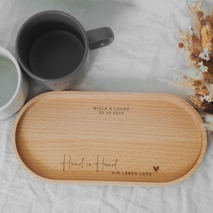 Pu&ograve; includere: Un vassoio di legno chiaro con bordi arrotondati, inciso con le parole "Hand in Hand" e "Ein Leben Lang". Due tazze, una grigia e una bianca, sono accanto al vassoio. Sullo sfondo una composizione di fiori secchi.