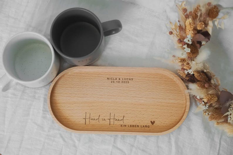 Pu&ograve; includere: Un vassoio di legno chiaro con bordi arrotondati, inciso con le parole "Hand in Hand" e "Ein Leben Lang". Due tazze, una grigia e una bianca, sono accanto al vassoio. Sullo sfondo una composizione di fiori secchi.