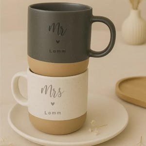 Pu&ograve; includere: Due tazze in ceramica impilate, una grigio scuro e una bianca, con base beige. La tazza grigio scuro &egrave; etichettata "Mr" con un cuore e il nome "Lomm". La tazza bianca &egrave; etichettata "Mrs" con un cuore e il nome "Lomm".