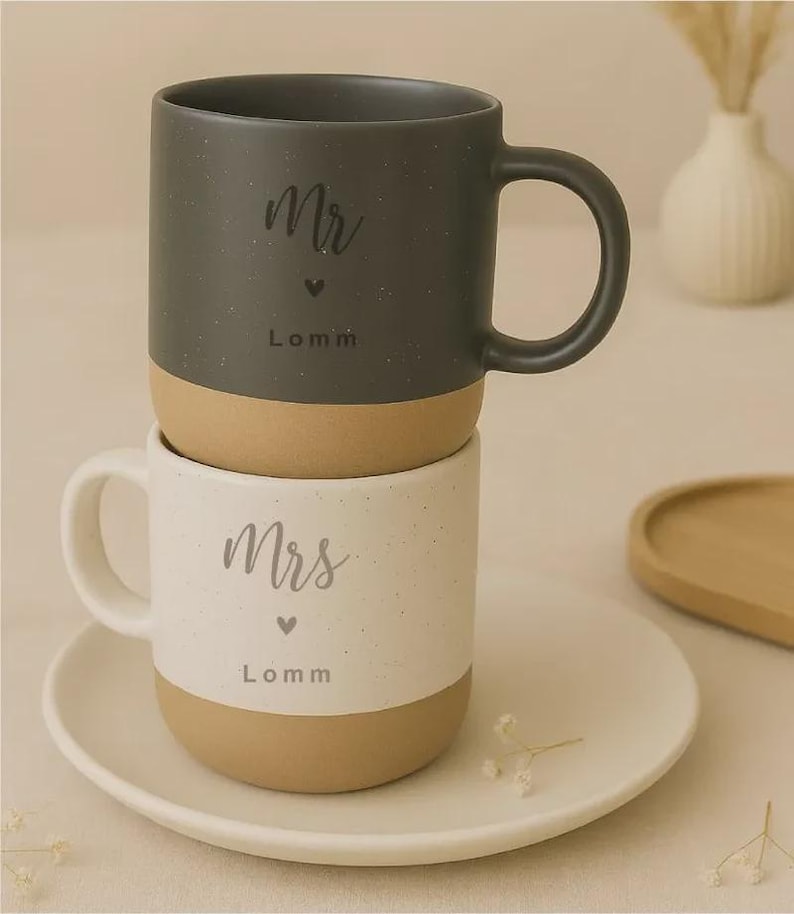 Pu&ograve; includere: Due tazze in ceramica impilate, una grigio scuro e una bianca, con base beige. La tazza grigio scuro &egrave; etichettata "Mr" con un cuore e il nome "Lomm". La tazza bianca &egrave; etichettata "Mrs" con un cuore e il nome "Lomm".
