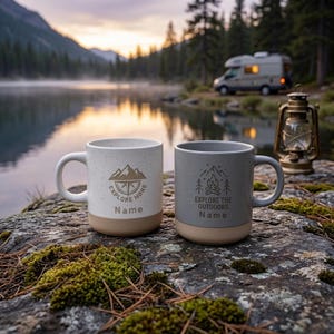 Könnte beinhalten: Zwei Keramikbecher mit gesprenkelter Oberfläche und einfarbigen Böden. Der weiße Becher zeigt ein Berggrafik-Design mit dem Text "EXPLORE MORE" und "Name". Der graue Becher zeigt ein Lagerfeuer-Motiv mit dem Text "EXPLORE THE OUTDOORS" und "Name".