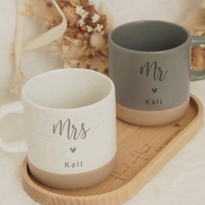 Könnte beinhalten: Zwei Keramikbecher, einer weiß und einer grau, stehen auf einem Holztablett. Der weiße Becher hat den Text "Mrs" und "Kall" mit einem Herz, während der graue Becher "Mr" und "Kall" mit einem Herz hat. Die Becher haben ein gesprenkeltes Design.