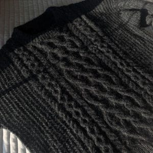 May include: A dark gray knitted sweater with a textured cable knit design on the front. The sweater has a crew neck and long sleeves. The knitwear appears soft and warm, suitable for cooler weather.
