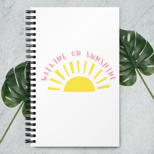 Puede incluir: Cuaderno blanco con espiral con la frase "WALKING ON SUNSHINE" en letras rosas sobre un sol amarillo. El cuaderno está rodeado de hojas tropicales verdes.