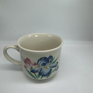 May include: Cream ceramic mug with a curved handle. The mug has a floral design featuring blue and pink iris flowers and green leaves. The mug is empty and sits on a white surface.