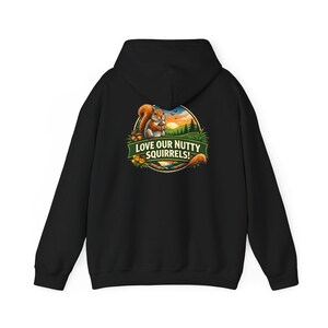 Puede incluir: Sudadera con capucha negra con un gráfico colorido de una ardilla sosteniendo una nuez, sobre un fondo de puesta de sol. El texto "LOVE OUR NUTTY SQUIRRELS!" se muestra en una pancarta verde. La sudadera tiene capucha y bolsillo de canguro.
