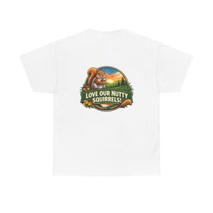 Puede incluir: Camiseta blanca con un diseño gráfico colorido de una ardilla sosteniendo una nuez, con el texto "LOVE OUR NUTTY SQUIRRELS!". El diseño incluye una puesta de sol, árboles y follaje, creando una ilustración con temática de la naturaleza.