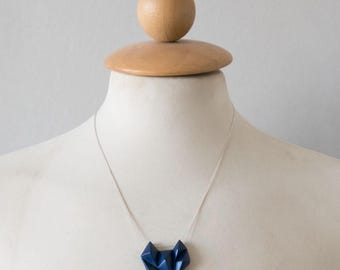 May include: A necklace featuring a dark blue, geometric, faceted pendant suspended from a thin, silver-toned metal chain. The pendant has a sharp, angular, multi-sided shape that hangs centrally from the delicate chain against a neutral display.