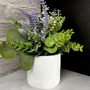 May include: A white ceramic pot with a textured, concentric circle design, filled with a mix of artificial greenery and lavender. The arrangement includes large, round green leaves and purple lavender sprigs, creating a fresh, natural look.