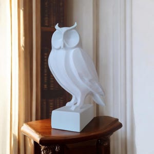 May include: A white owl sculpture with a geometric design stands on a rectangular base. The owl is perched on a wooden table with a curved edge. The background includes a bookshelf and a curtain.