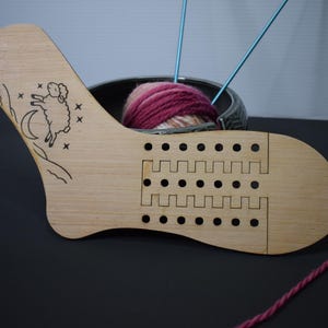 May include: A wooden sock knitting gauge with a sheep and moon design. The gauge has multiple holes and cutouts for measuring. A ball of pink and white yarn sits in a metal bowl behind the gauge, with two blue knitting needles.