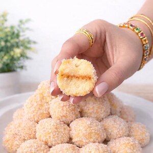 Peut inclure: Une assiette de biscuits ronds et clairs, certains avec un enrobage texturé. Un biscuit est tenu, révélant une garniture. Les biscuits sont disposés sur une assiette blanche, avec une plante en arrière-plan.