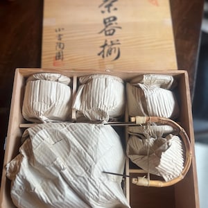 May include: A wooden tea set box with Japanese characters on the lid. Inside, the box contains wrapped tea cups and a bamboo tea whisk. The tea set is presented in a light brown wooden box.