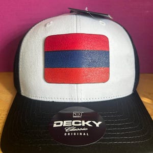 May include: A trucker hat with a white front panel and a black mesh back. The hat features a square patch with red, blue, and red stripes. The hat has a black brim and a "DECKY Classic Original" sticker.