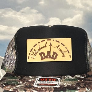 May include: Black and camouflage trucker hat with a mesh back and a curved brim. The hat features a rectangular patch with the words "Oldschool Analog Dad" in a stylized font. The hat has a snap closure.