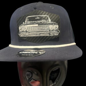 May include: A navy blue snapback cap featuring a white rope detail across the base of the crown. A black carbon fibre-style patch on the front displays a white line-art illustration of a classic car. A black sticker with the text DECKY ORIGINAL is visible.