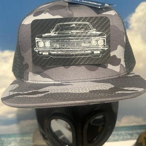 May include: A grey camouflage trucker hat featuring a rectangular black patch with a white line art illustration of the front of a classic car. The hat has a flat brim and a mesh back, with a small blue tag attached to the top of the crown.