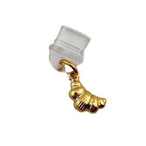May include: A gold-colored charm in the shape of a croissant hangs from a small gold ring attached to a clear earring back. The croissant charm has detailed ridges and a shiny finish, set against a white background.