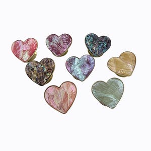 May include: A collection of eight heart-shaped phone grips featuring iridescent shell inlays. Each piece displays unique patterns and colors, including shades of pink, purple, blue, green, and gold, all framed with a metallic gold-toned edge.