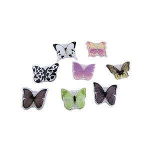 May include: A collection of eight butterfly-shaped decorative items featuring various patterns and colors, including a black and white cow print, a solid purple, a light green, a dark grey, a yellow-toned wing design, and a multi-colored pastel wing.