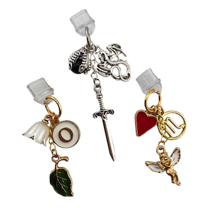 May include: Three decorative earring charms with various designs. One charm features a dragon, a black gem, and a sword. Another has a white flower, a white and gold circle, and a green leaf. The third has a red heart, a gold angel, and a gold zodiac symbol.