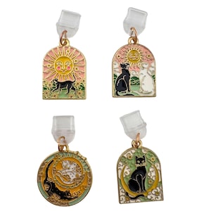 May include: Four pairs of gold-toned cat-themed earrings. Each earring features a different design: a black cat with a sun, two cats with a sun, a cat on a crescent moon, and a cat on a crescent moon. The earrings have a post back.