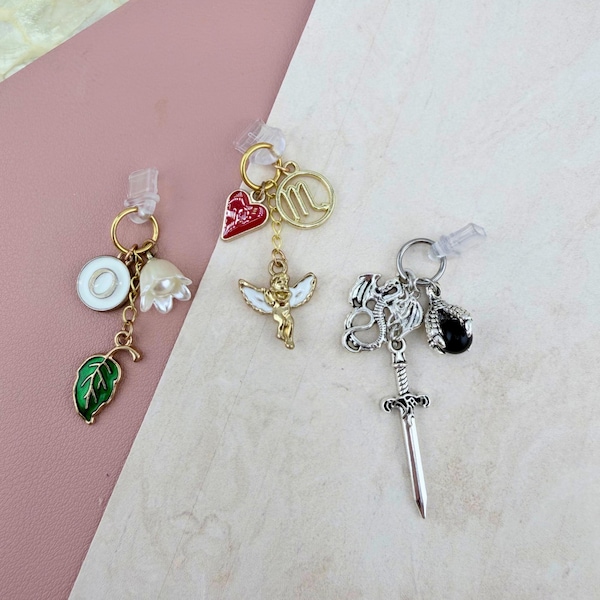 Puede incluir: Tres dijes decorativos unidos a anillos de plástico transparente. El primero presenta un círculo blanco con la letra O, una flor en forma de campana y una hoja verde. El segundo tiene un corazón rojo, un símbolo de Escorpio dorado y una figura alada. El tercero tiene un dragón plateado, una espada y una garra negra.