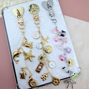 May include: Three decorative keychains with metal clasps. The first features gold-toned sea life charms including a crab, dolphin, starfish, turtle, and shell. The second has gold-toned charms like a letter L, a cross, and a sunflower. The third has colorful character charms.