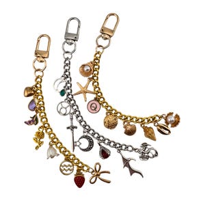 Pode incluir: Três pingentes de corrente decorativos com fechos de lagosta. Um é dourado, um prateado e um dourado. Cada corrente apresenta vários pingentes, incluindo um morango, uma espada, uma concha, uma estrela do mar e uma pérola.
