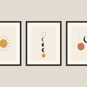 Op de afbeelding: Een set van drie ingelijste wandkunstprints met zwarte lijsten. De eerste print toont een gele zon met stralen. De tweede toont een verticale reeks maanfasen. De derde toont een maansikkel en een terracotta cirkel met kleine sterren.