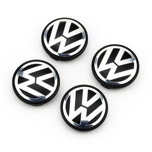 Può includere: Quattro coprimozzi neri e argentati con il logo Volkswagen. I coprimozzi sono rotondi con sfondo nero e un emblema VW argentato al centro. Sono disposti a quadrato su sfondo bianco.