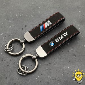 May include: Two black leather keychains with silver hardware. One keychain features the BMW logo and the word "BMW" in white. The other keychain has the BMW M logo. Both keychains have red stitching.