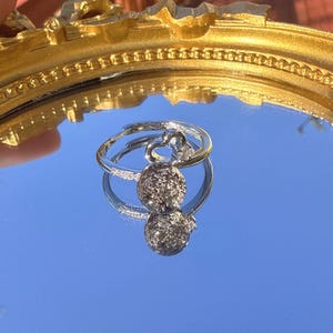 May include: A silver ring with a double-sphere pendant. The ring is set against a reflective blue surface, with a gold-colored ornate frame in the background. The pendant has a textured, speckled appearance.