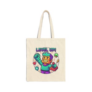 Puede incluir: Bolso tote de lona beige con un gráfico colorido de un personaje pixelado sosteniendo un joystick, con las palabras "LEVEL UP!". El diseño incluye corazones, un hongo y rayos, evocando un tema de videojuego.