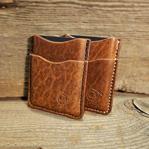 May include: Two brown leather card holders with a distressed finish. Each wallet has multiple pockets and is stitched with black thread. The wallets are embossed with a logo and sit on a wooden surface.