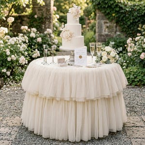 May include: A tiered wedding cake with floral decorations sits on a round table covered in a cream-colored, ruffled fabric. Champagne flutes, a bouquet, and a ring box are also on the table. The scene is set outdoors with greenery in the background.
