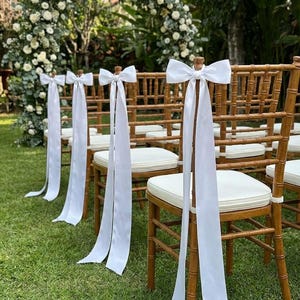 Peut inclure: Rangée de chaises en bambou avec coussins blancs et nœuds en satin blanc. Les chaises sont disposées sur une pelouse verte, avec une arche florale en arrière-plan, créant un cadre de mariage ou d'événement.