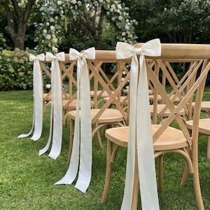 Op de afbeelding: Houten stoelen met crèmekleurige strikken en lange linten staan op een grasveld. De stoelen zijn opgesteld voor een buitenevenement, met een bloemenboog op de achtergrond. De strikken en linten voegen een vleugje elegantie toe.