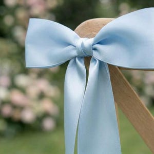 Puede incluir: Un lazo de satén azul claro atado a una silla de madera. El lazo está centrado y tiene largas cintas que fluyen. El fondo está borroso, mostrando vegetación y colores pastel suaves, lo que sugiere un evento al aire libre.