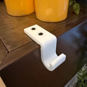 May include: A white, plastic shelf bracket with a curved hook design. The bracket has two black screws and is designed to attach to a shelf or other surface. The bracket is shown on a dark brown surface.