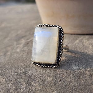 May include: A silver ring featuring a large, rectangular moonstone. The moonstone has a pearly white color with subtle blue and yellow iridescence. The ring's setting is silver with a twisted rope design around the stone's edge.