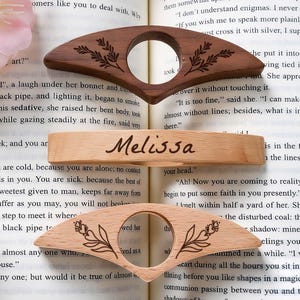 May include: Three wooden book page holders with floral designs and a personalized bookmark with the name "Melissa" are displayed on an open book. The holders are made of light and dark wood.