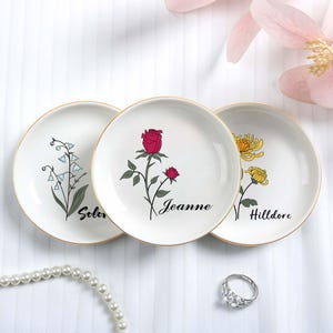 Puede incluir: Tres platos decorativos de cerámica blanca con bordes dorados, cada uno con un diseño floral diferente: lirios de los valles, una rosa roja y crisantemos amarillos. El nombre "Jeanne" está escrito en un plato.