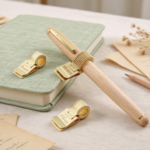 May include: Three gold-colored metal pen clips with spring-loaded loops, each engraved with a name: Bonnie, Henry, and Olivia. One clip holds a light-colored wooden pen. The clips feature a flat base and a coiled wire loop for holding pens.