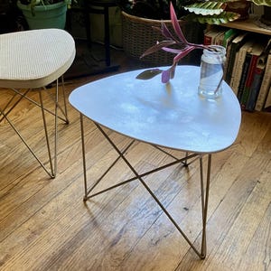 May include: A white, teardrop-shaped side table with gold-colored wire legs. A small glass jar with water and a purple plant stem sits on the table. Another stool with a white seat is in the background. The room is filled with potted plants.