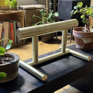 May include: A gold-toned metal display stand featuring a horizontal cylindrical bar supported by two vertical posts connected to a base composed of two parallel horizontal cylindrical bars, creating a sturdy H-shaped frame structure.